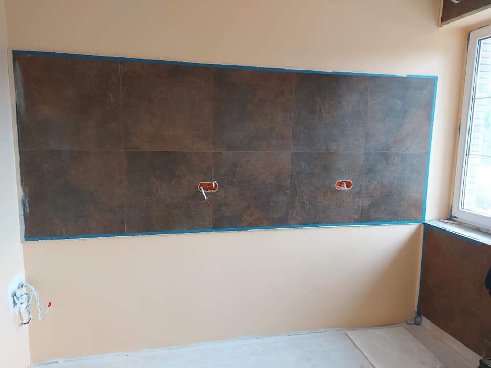 Ściana w trakcie wykańczania, z zamontowanymi brązowymi płytkami imitującymi kamień, zabezpieczonymi niebieską taśmą malarską, widoczne wystające kable elektryczne, ściany pomalowane na beżowo.