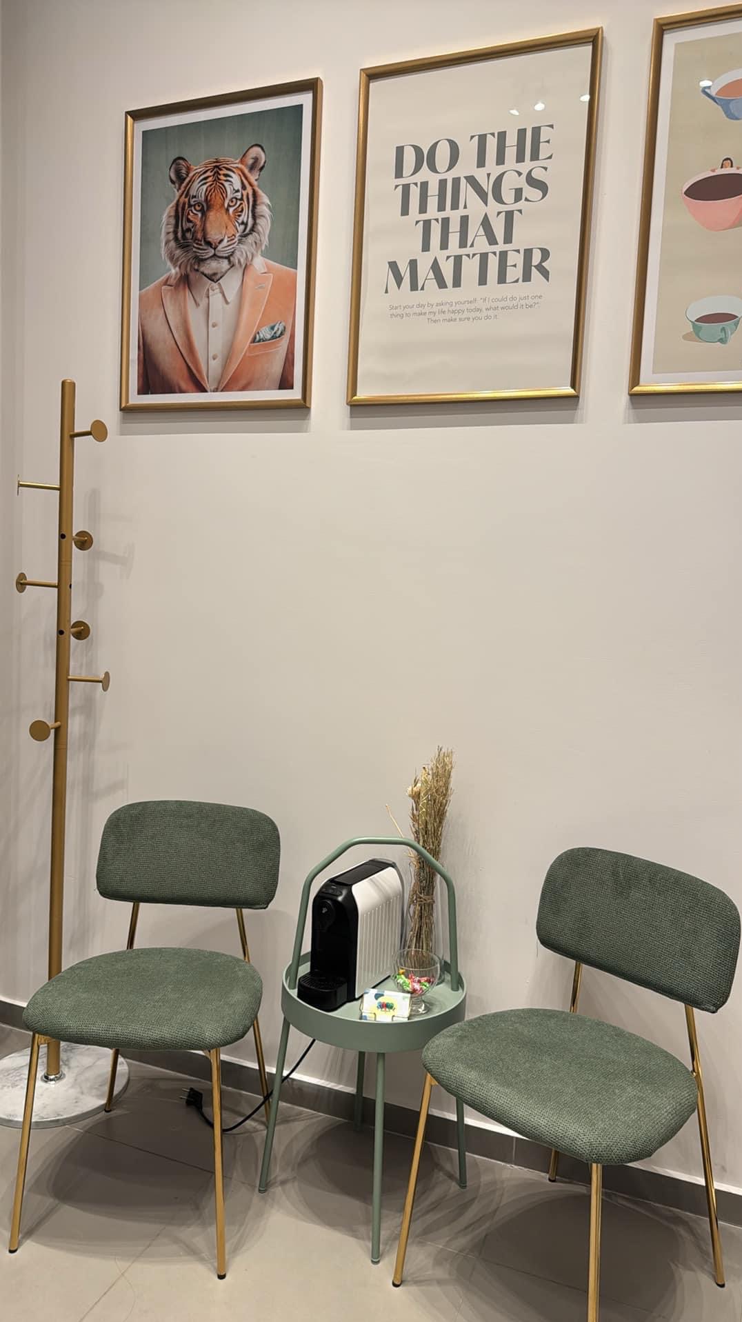 Minimalistyczna poczekalnia z dwoma krzesłami w odcieniach zieleni, stolikiem z ekspresem do kawy i dekoracyjnymi obrazami w złotych ramach na ścianie.
