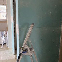 Maciej Siwiec - Ściana z płyt gipsowo-kartonowych w trakcie montażu, widoczna drabina malarska i fragmenty konstrukcji. Surowy stan wykończenia wnętrza, widok na okno w tle.