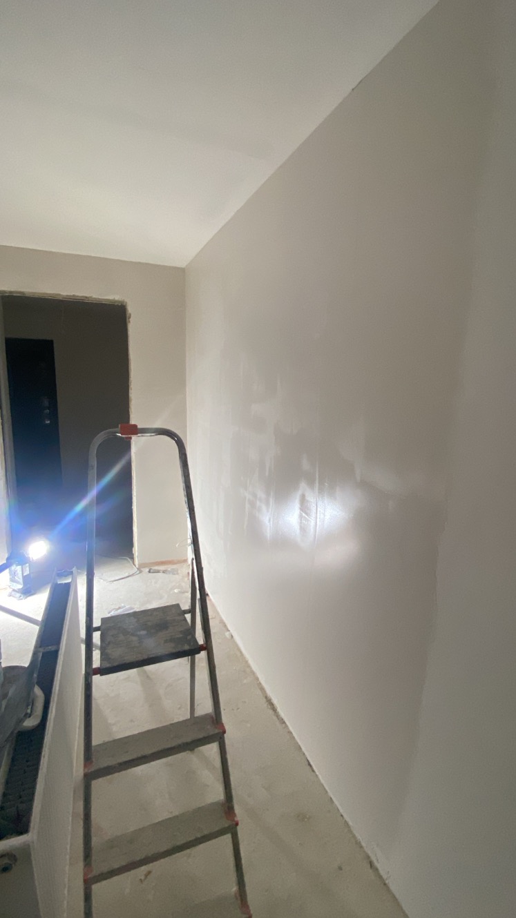 Świeżo pomalowana, gładka ściana w jasnym odcieniu, z widoczną drabiną i oświetleniem roboczym. Widoczny fragment sufitu i podłogi w trakcie prac wykończeniowych.