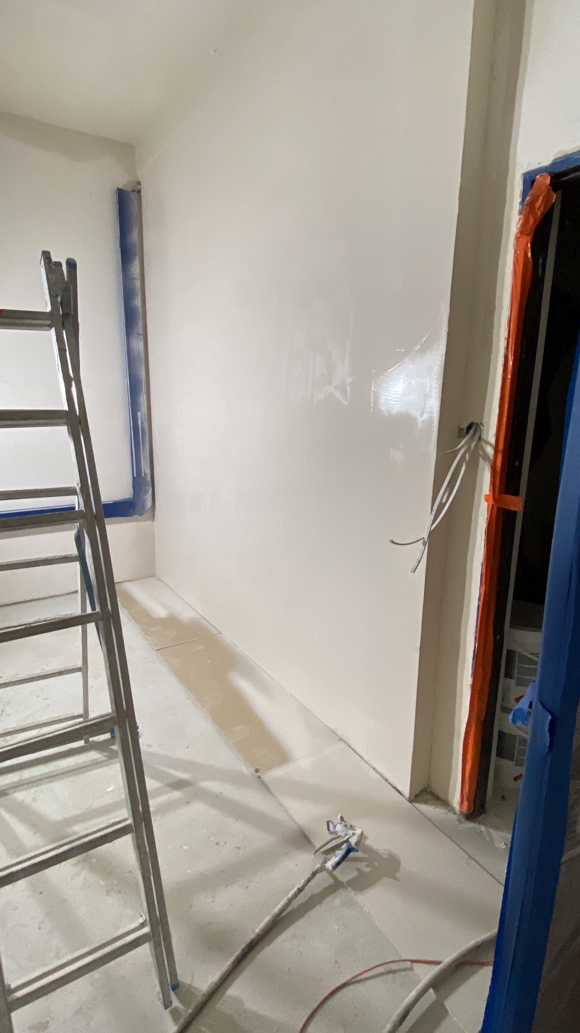 Świeżo położona, gładka, jasna gładź na ścianie w trakcie remontu. Widoczna drabina, taśmy malarskie i pistolet natryskowy do gładzi. Ujęcie z bliska, wnętrze w stanie surowym.