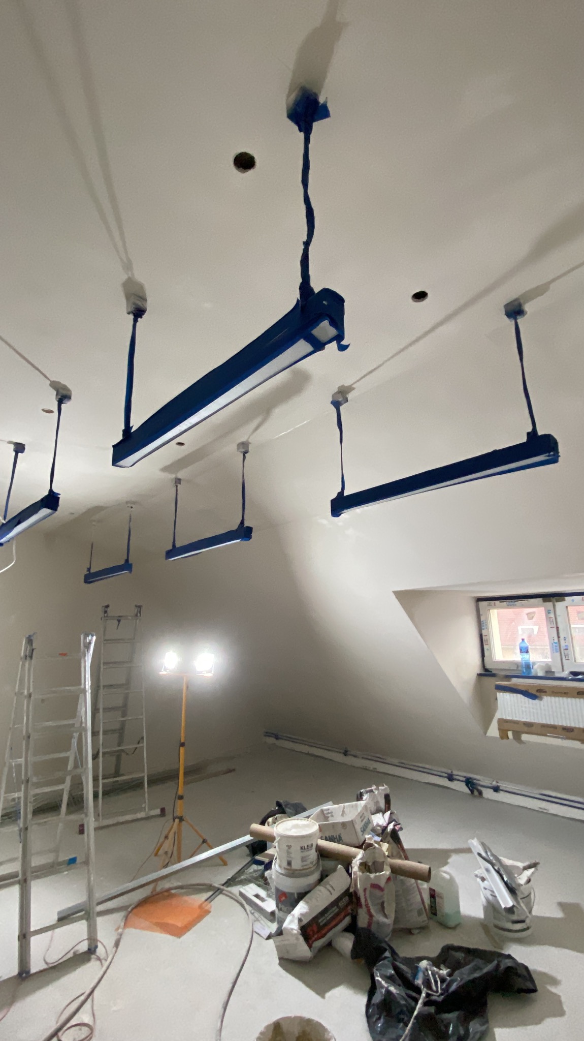 Przygotowania do malowania poddasza: lampy zabezpieczone taśmą, drabiny, worki z materiałami budowlanymi, okno w skosie. Widok na sufit i część pomieszczenia.