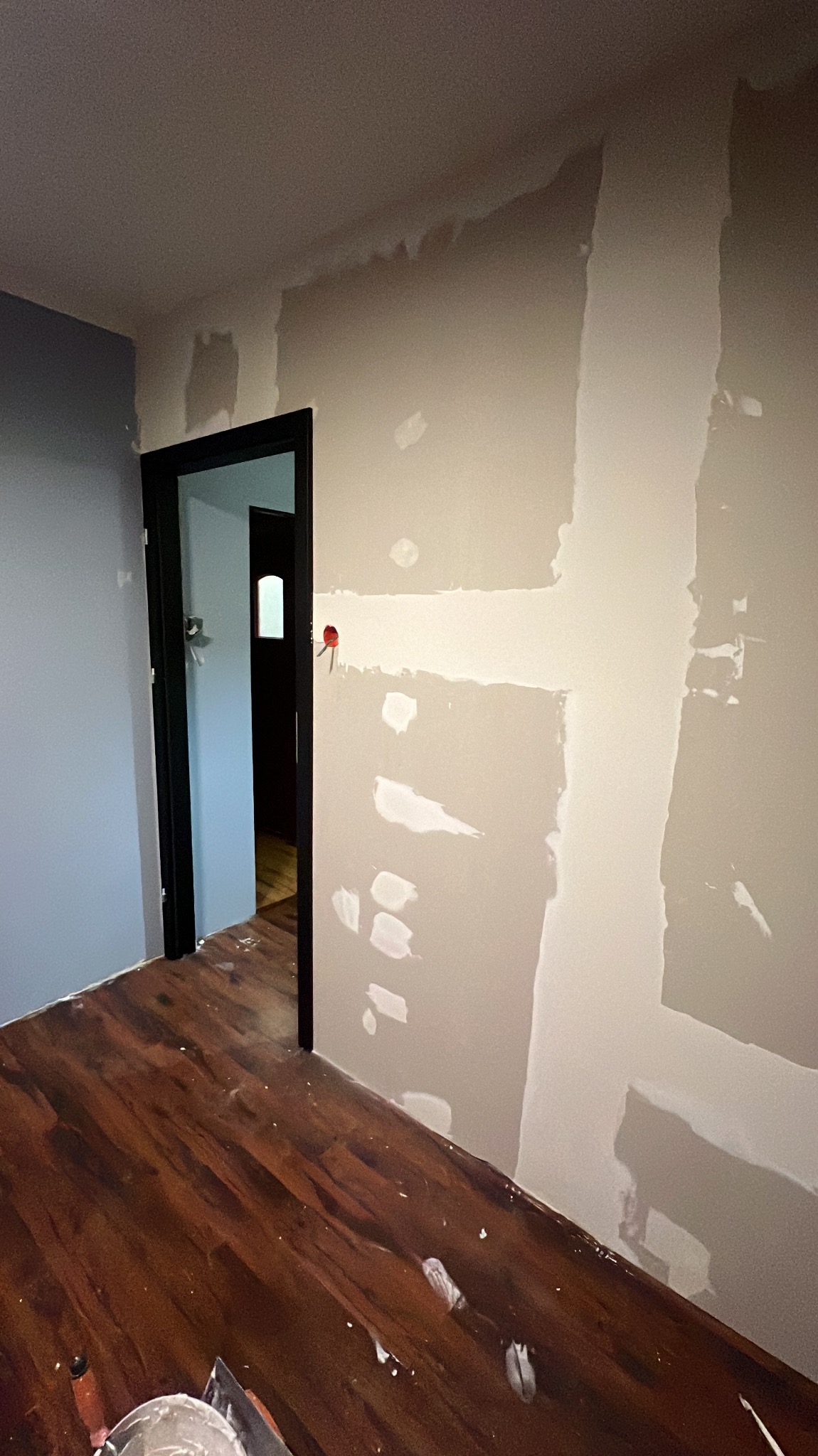 Pokój w trakcie szpachlowania: fragment ściany z nałożoną gładzią, widoczne narzędzia na podłodze z paneli, czarna futryna drzwi. Prace wykończeniowe.
