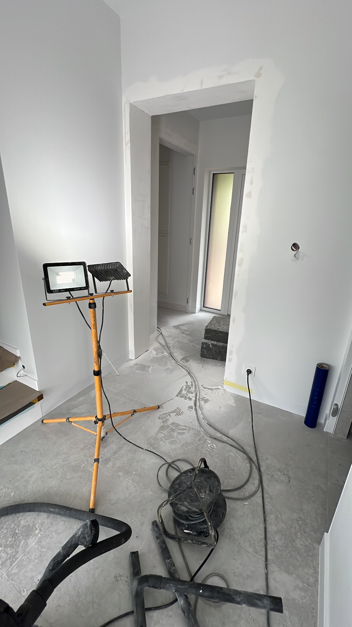 Wnętrze w trakcie szpachlowania: widoczne ślady gładzi na ścianach, lampa budowlana na statywie, przewody i osprzęt budowlany na podłodze. Otwór drzwiowy w tle.