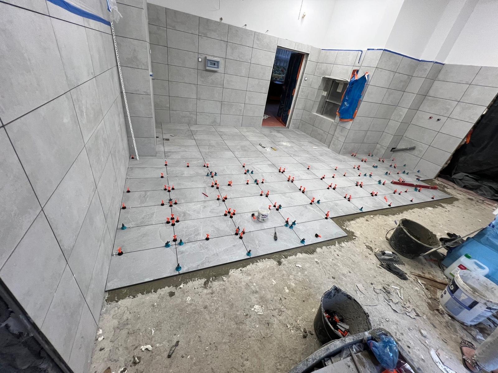 Układanie szarych płytek w łazience z użyciem systemu poziomowania. Widoczne krzyżyki i kliny. Częściowo wykończona podłoga i ściany. W tle otwarte drzwi.