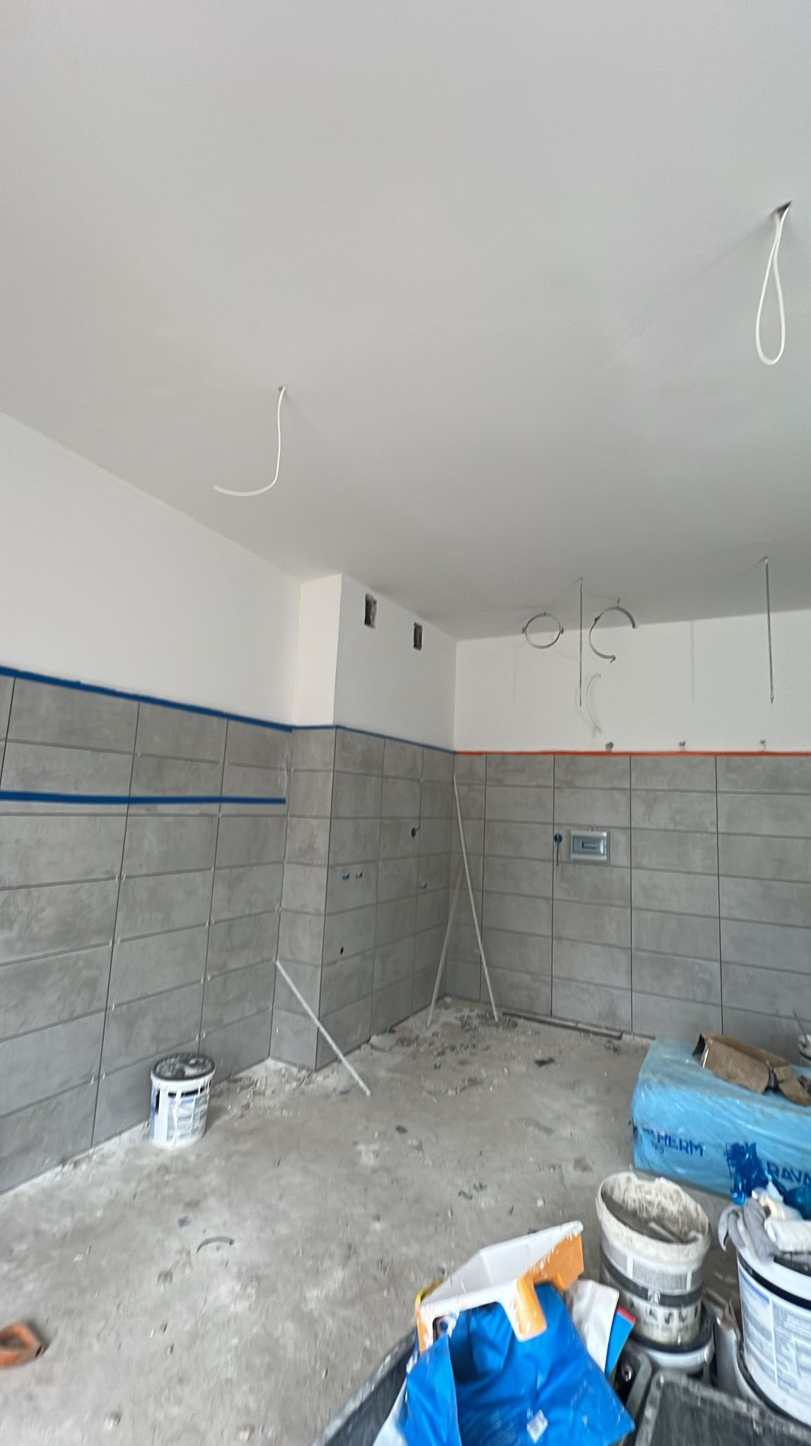 Wnętrze łazienki w trakcie prac glazurniczych: szare płytki ułożone do połowy ściany, widoczne przewody, narzędzia i materiały budowlane. Niebieska taśma malarska wyznacza górną krawędź płytek.