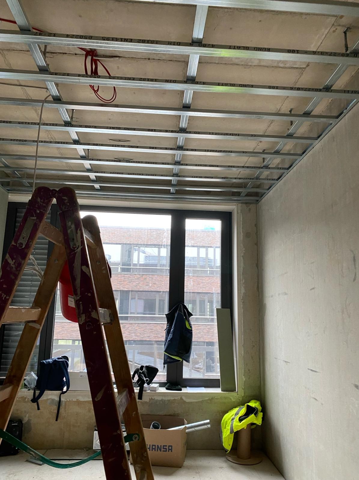 Montaż sufitu podwieszanego z płyt karton-gips, widoczna metalowa konstrukcja, drabina malarska i okno z widokiem na budynek. Wnętrze w trakcie prac wykończeniowych.