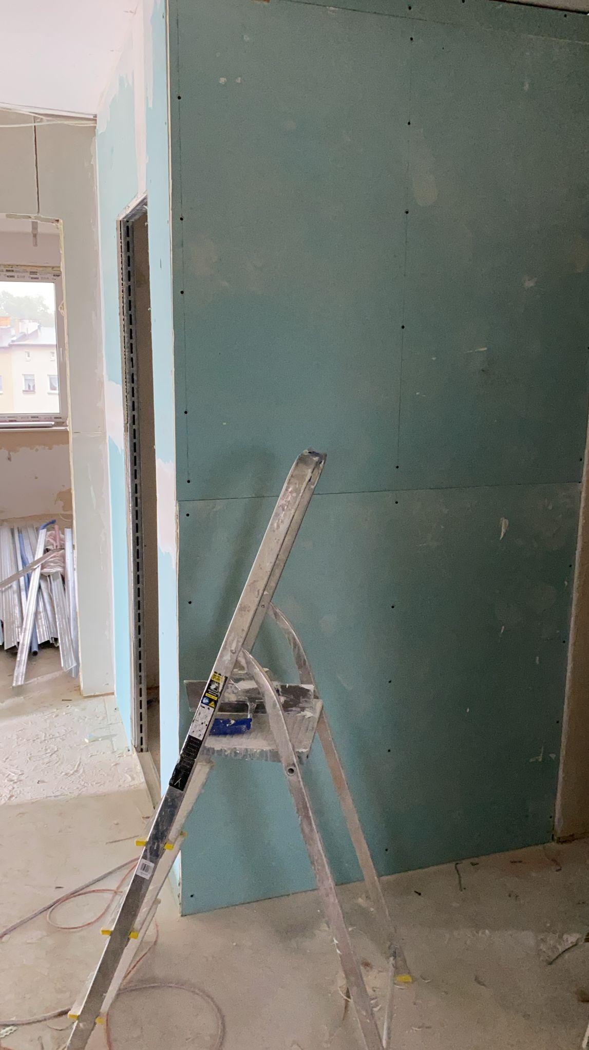 Ściana z płyt gipsowo-kartonowych w trakcie montażu, widoczna drabina malarska i fragmenty konstrukcji. Surowy stan wykończenia wnętrza, widok na okno w tle.