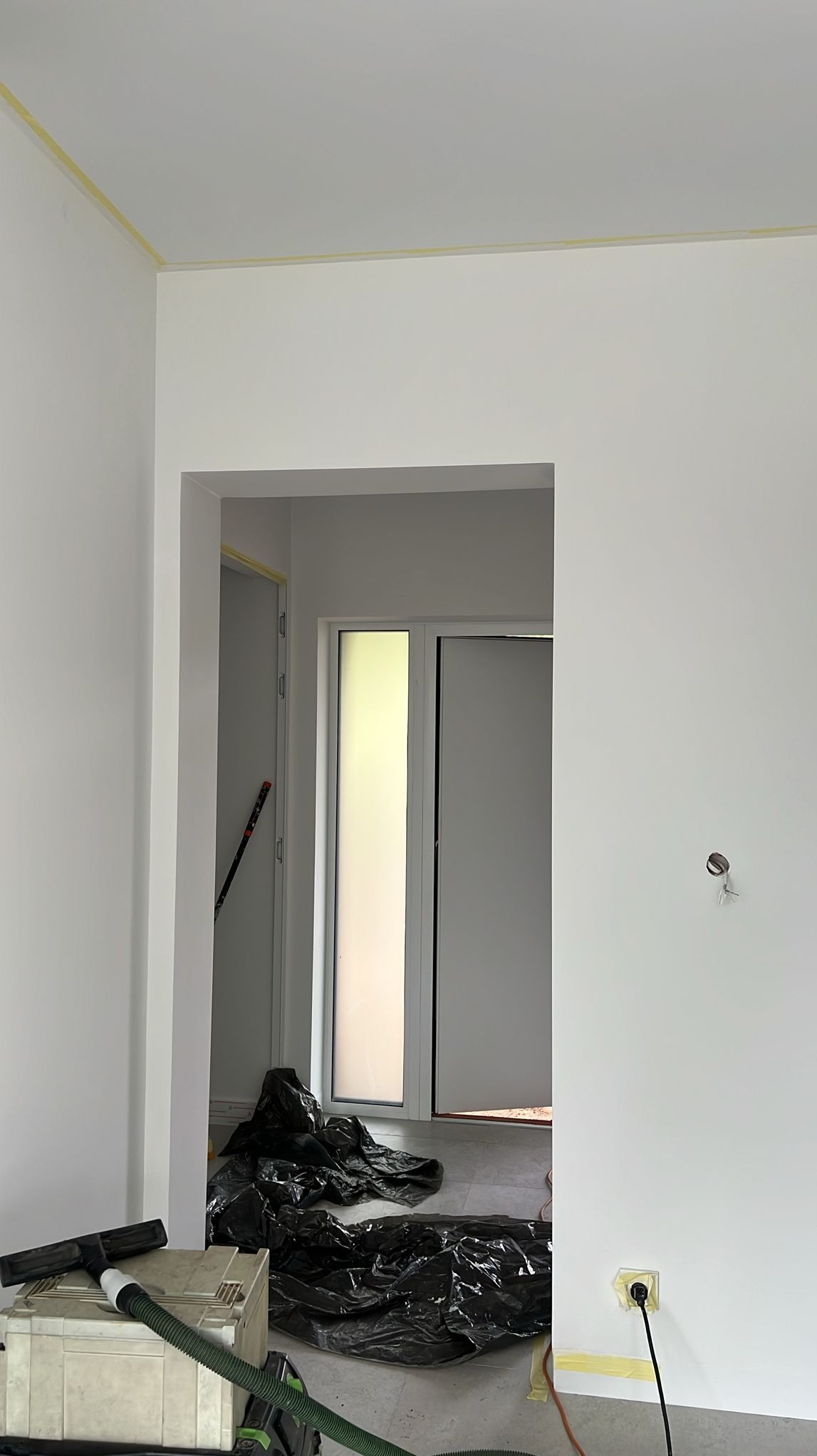 Białe ściany po malowaniu, zabezpieczone taśmą malarską przy suficie i gniazdkach. Widoczny odkurzacz przemysłowy i folia ochronna na podłodze.