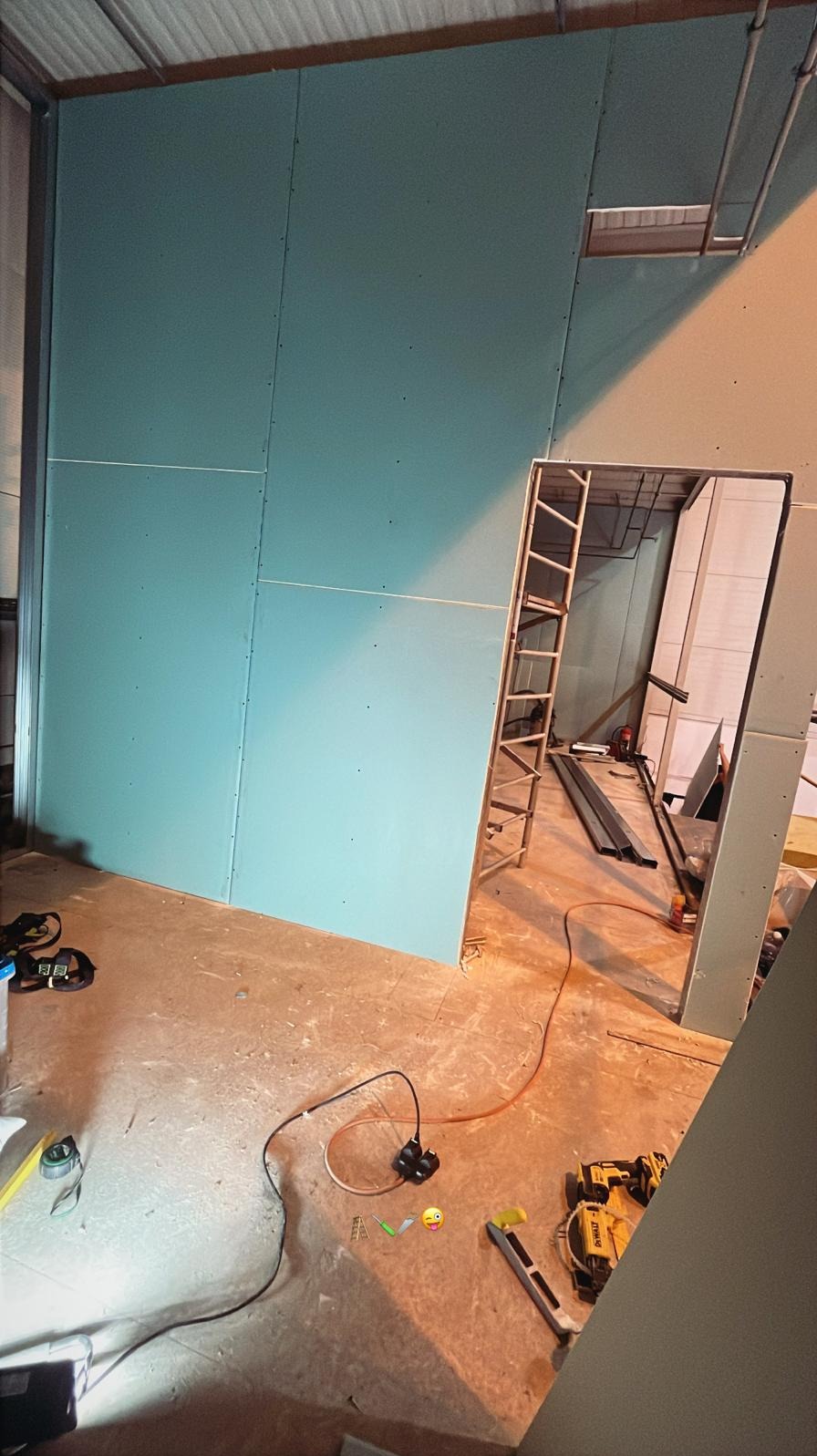 Montaż płyty karton gips na ścianie z widocznym otworem drzwiowym i narzędziami na podłodze. Widoczna drabina oparta o ścianę.