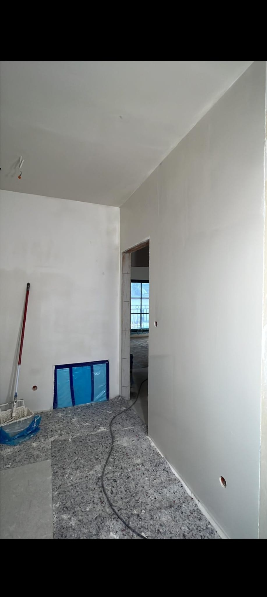 Wnętrze w trakcie prac wykończeniowych: gładkie, białe ściany, zabezpieczone okno, narzędzia malarskie i kabel na podłodze. Widok na dalsze pomieszczenia.