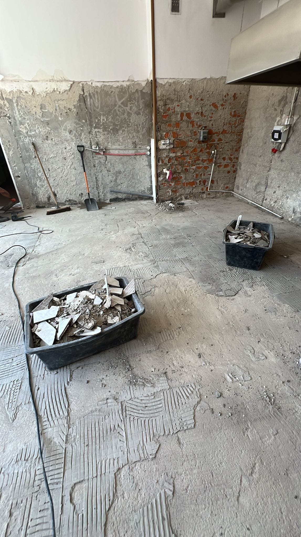 Demontaż płytek podłogowych w trakcie remontu kuchni. Widoczne resztki kleju, gruz w pojemnikach i fragmenty instalacji. Ściany częściowo otynkowane, częściowo z cegły.