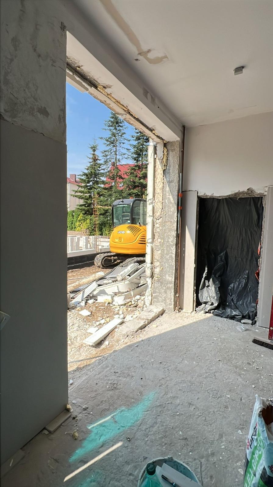 Wnętrze w trakcie remontu z widokiem na żółtą koparkę przez otwór w ścianie. Gruz i materiały budowlane na podłodze. Otwór drzwiowy zabezpieczony folią.