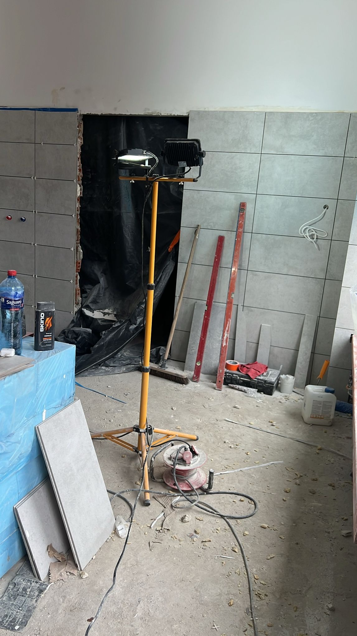 Ściana wyłożona szarymi płytkami w trakcie prac glazurniczych, widoczna lampa budowlana, poziomice i narzędzia. Fragment otworu zabezpieczony folią.