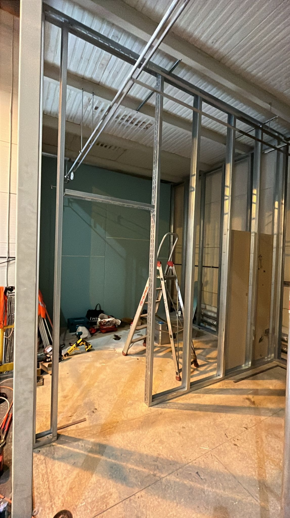 Metalowa konstrukcja ścian działowych z płyt karton-gips, widoczna drabina i narzędzia, surowy stan wnętrza w trakcie prac wykończeniowych.