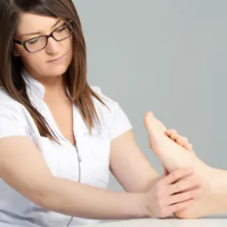 Kobieta w białym uniformie i okularach delikatnie masuje stopę pacjenta na białym stole, na jasnoszarym tle.