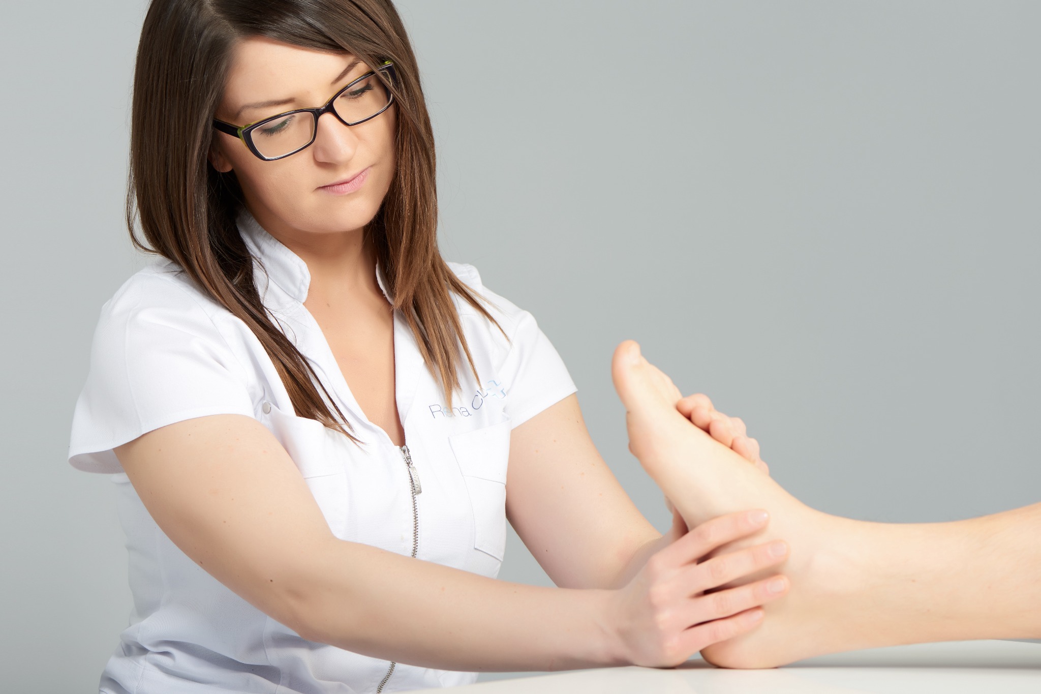 Kobieta w białym uniformie i okularach delikatnie masuje stopę pacjenta na białym stole, na jasnoszarym tle.