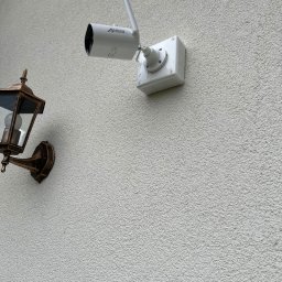 NKTelektrick - Biała kamera monitoringu marki Anran zamontowana na elewacji budynku obok stylizowanej, metalowej lampy zewnętrznej w kolorze starego złota.