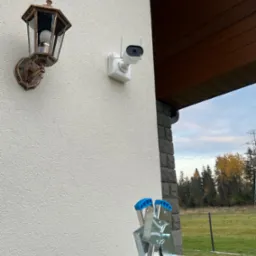 Biała kamera monitoringu z antenami zamontowana na elewacji domu obok ozdobnej lampy zewnętrznej; drabina aluminiowa oparta o ścianę, widok z dołu.