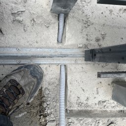 NKTelektrick - Szare, karbowane rury ochronne ułożone w wykopie pod szyną bramy przesuwnej, widoczny but roboczy dla skali.