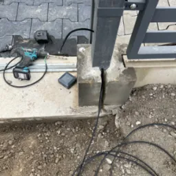 Montaż słupka bramy z ukrytym przewodem elektrycznym, wiertarka akumulatorowa z wiertłem do betonu, zwój kabla na ziemi, kostka brukowa w tle.