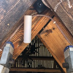 NKTelektrick - Widok z dołu na zamontowany czujnik dymu z rurką, przymocowany do drewnianej konstrukcji dachu. W tle widoczne prześwity desek i fragment drabiny.