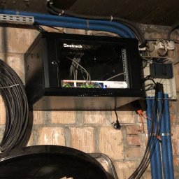 NKTelektrick - Zamontowana szafa rack Netrack na ceglanej ścianie z widocznymi kablami i elementami instalacji elektrycznej. Szafa częściowo otwarta, widoczne urządzenia sieciowe.