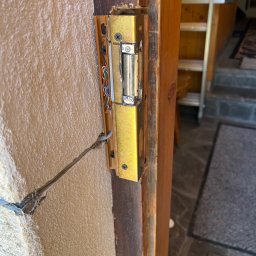 NKTelektrick - Złoty elektrozaczep zamontowany na drewnianej futrynie drzwi, z widocznymi przewodami i fragmentem ściany o nierównej fakturze. Widoczna drabina w tle.