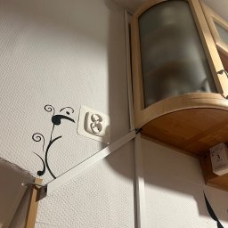 NKTelektrick - Białe gniazdko elektryczne na ścianie z dekoracyjną naklejką, widoczne elementy szafki wiszącej. Ujęcie z dołu, wnętrze mieszkania.