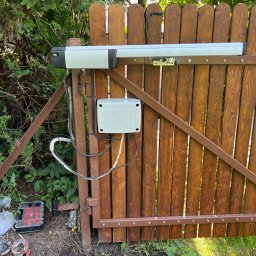 NKTelektrick - Brązowa brama z zamontowaną automatyką Somfy, widoczne okablowanie i skrzynka sterownicza. Tło: zieleń drzew i krzewów, narzędzia na ziemi.