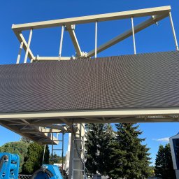 NKTelektrick - Zewnętrzny ekran LED na konstrukcji stalowej, widok z dołu na tle błękitnego nieba i zieleni drzew. Widoczny fragment podnośnika koszowego.