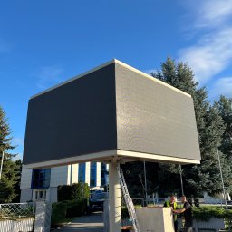 NKTelektrick - Wysoki ekran LED na konstrukcji, montowany przez dwóch mężczyzn w kamizelkach odblaskowych. W tle budynek z niebieskimi oknami i drzewa, błękitne niebo.