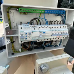 Otwarta skrzynka elektryczna z bezpiecznikami Hager, widoczne przewody w kolorach: żółto-zielonym, brązowym, niebieskim i szarym. Element instalacji zamontowany na konstrukcji.