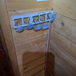 NKTelektrick - Rozdzielnica elektryczna z przewodami w drewnianej obudowie. Widoczne kable w kolorach: brązowym, niebieskim, żółto-zielonym. Częściowo zamontowane gniazdka.