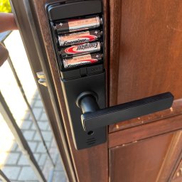 NKTelektrick - Czarny, nowoczesny zamek elektroniczny z klawiaturą numeryczną i uchwytem na baterie AA, zamontowany na drewnianych drzwiach wejściowych.