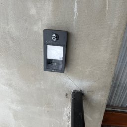 NKTelektrick - Zamontowany, czarny wideodomofon HiLook na szarej ścianie przy drzwiach. Widoczny fragment rury. Ujęcie z dołu, podkreślające nowoczesny design urządzenia.