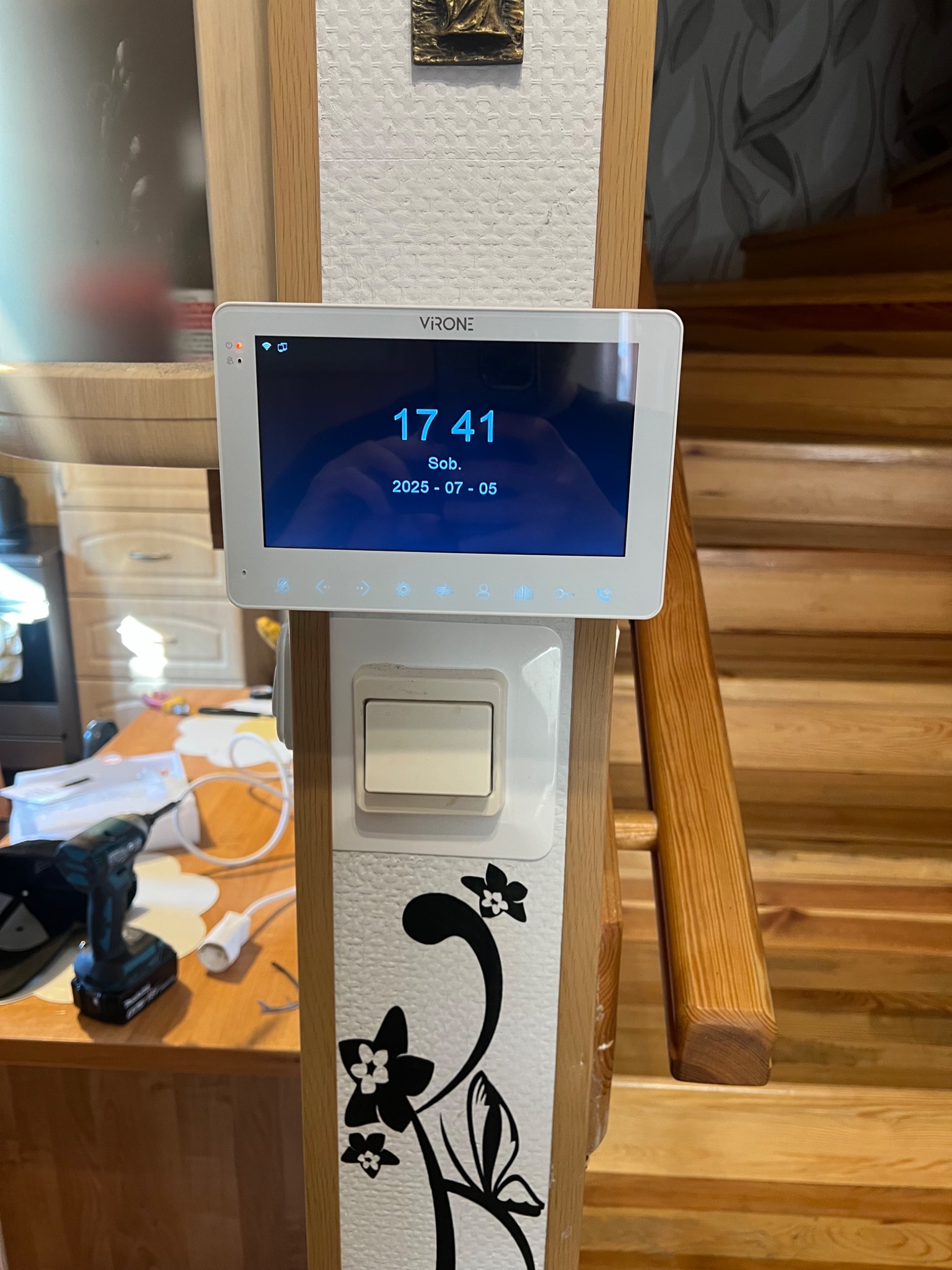Biały panel wideodomofonu Viron zamontowany na zdobionej kolumnie, z widocznym ekranem wskazującym godzinę i datę, w tle schody i meble.
