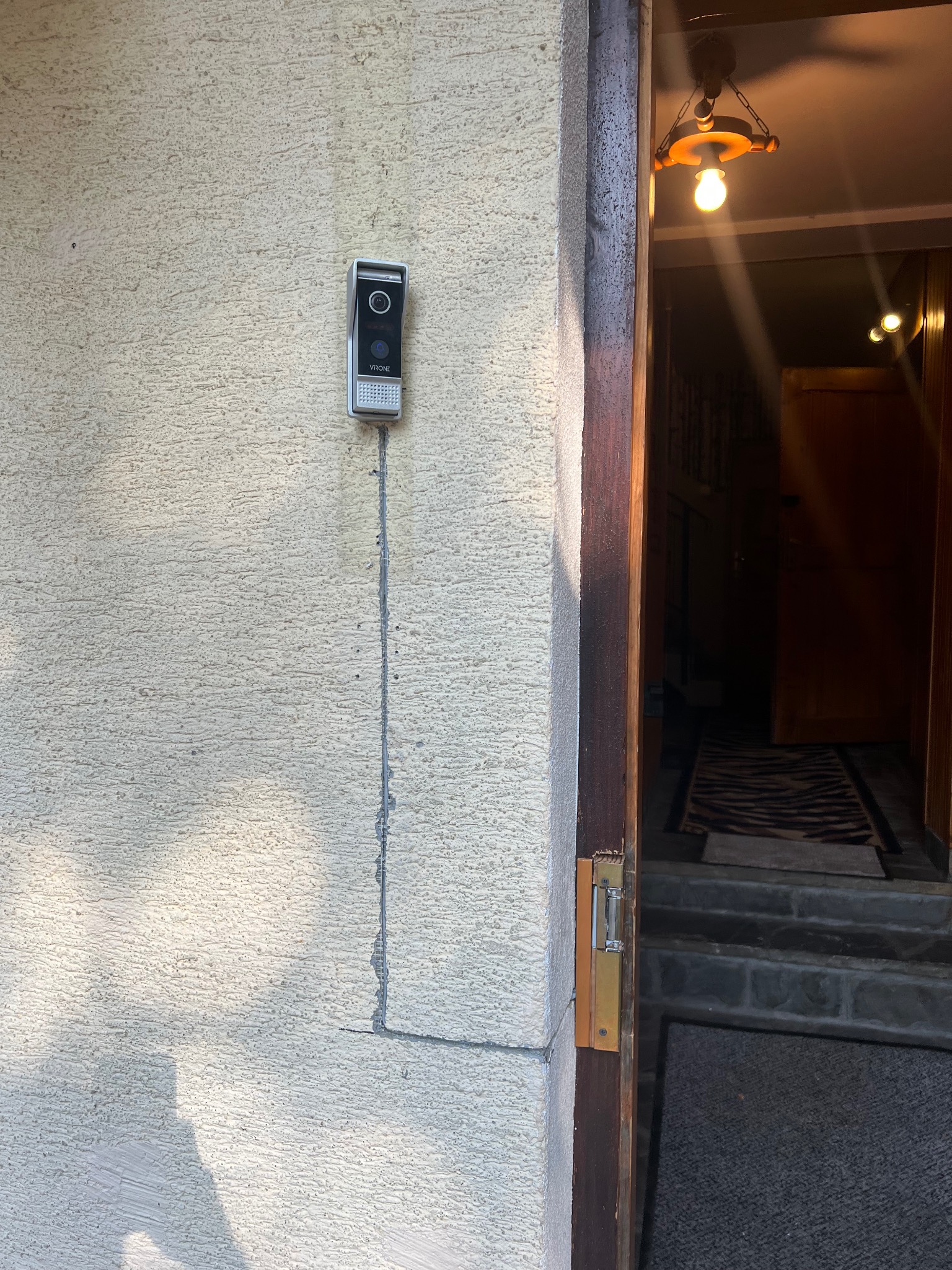 Zamontowany wideodomofon VRONE na teksturowanej ścianie zewnętrznej, widoczny kabel. Otwarte drzwi z widokiem na wnętrze domu i oświetlenie.
