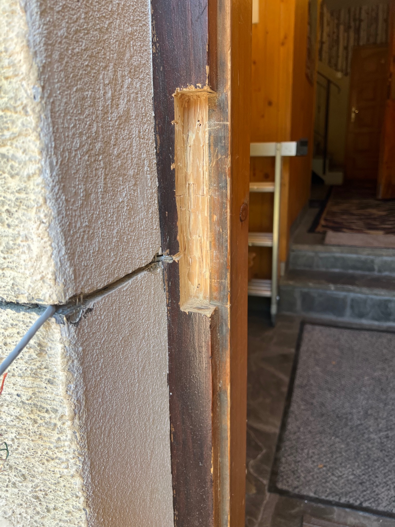 Drewniana futryna z wyżłobionym otworem na zamek, widoczne przewody elektryczne w ścianie. W tle fragment wnętrza domu z schodami i dywanem.
