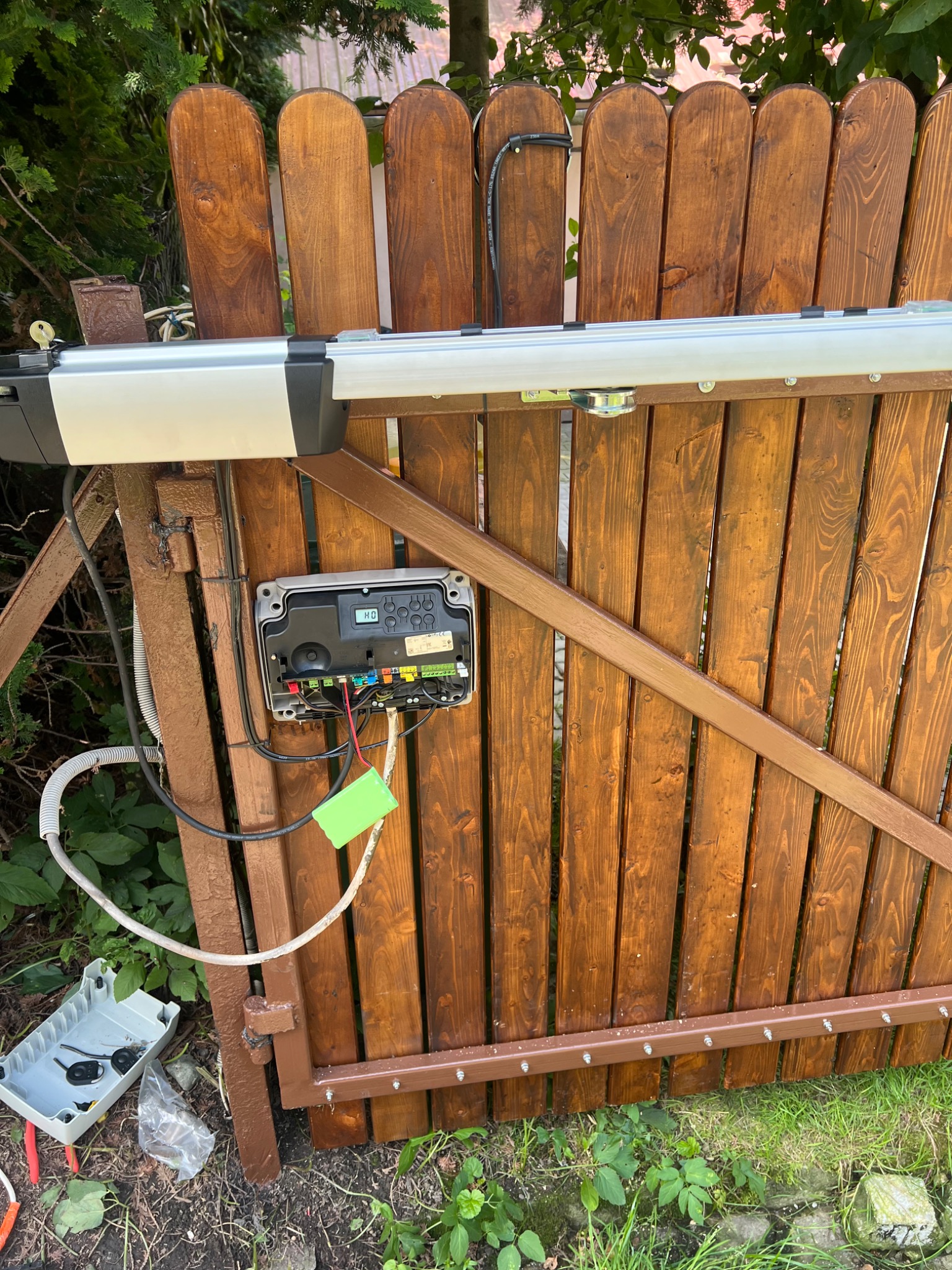 Drewniana brama z zamontowaną automatyką, widoczny sterownik z okablowaniem i mechanizm napędowy. Elementy montażowe leżą na ziemi obok bramy.