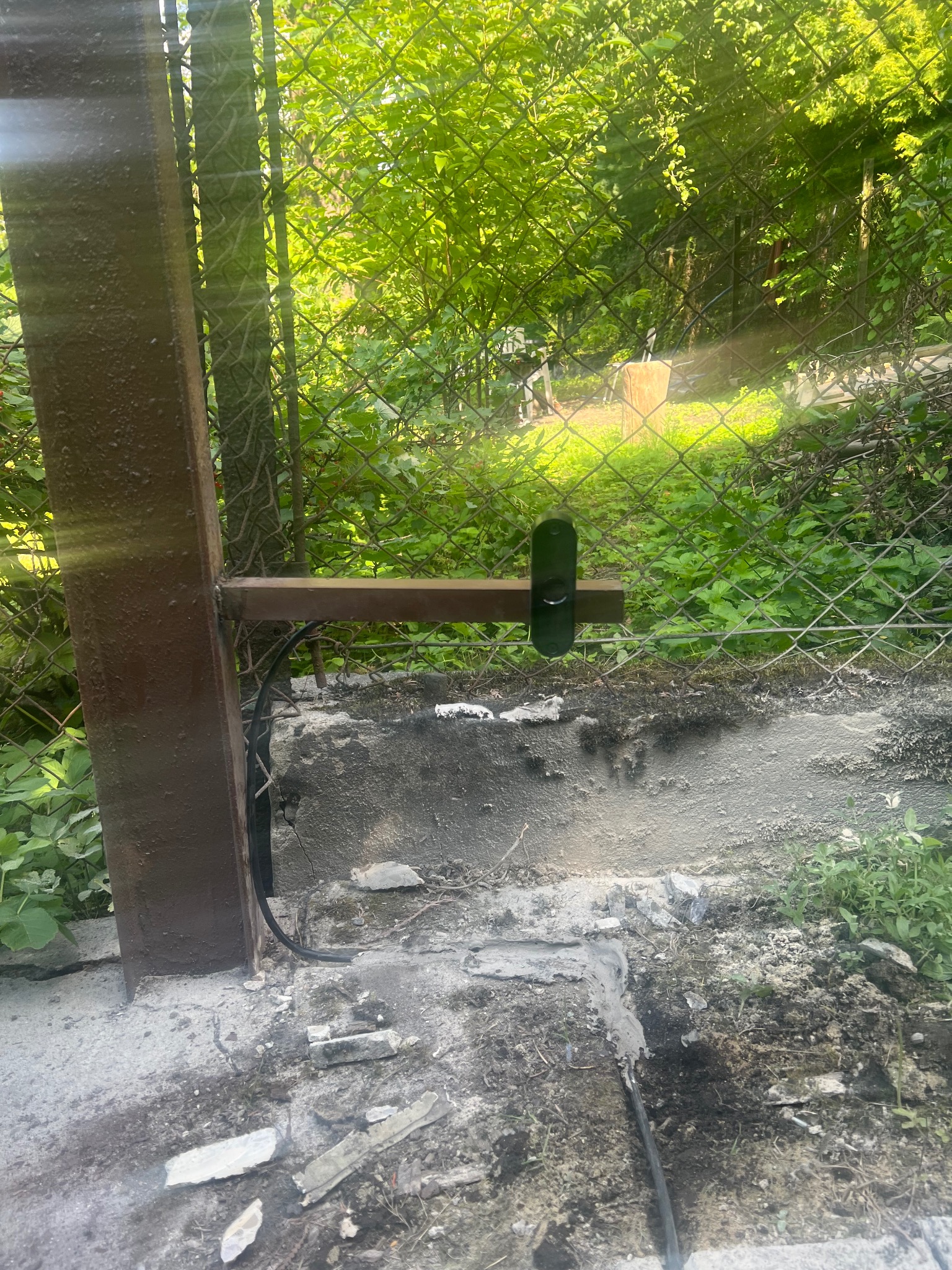 Zewnętrzny montaż wideodomofonu na metalowej bramie z siatki, widoczne przewody i elementy mocujące. Tło: zieleń. Kąt ujęcia z dołu.