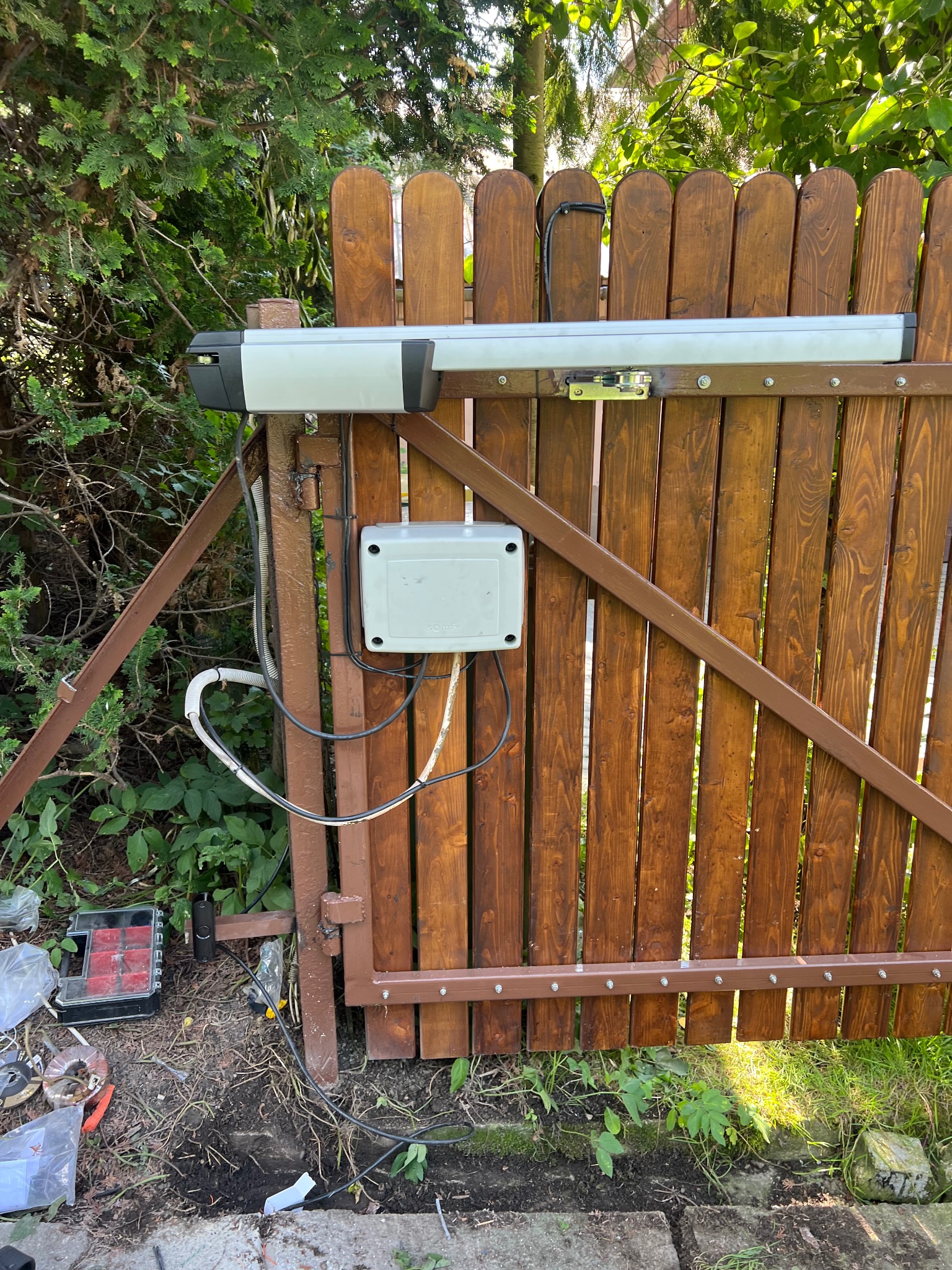 Brązowa brama z zamontowaną automatyką Somfy, widoczne okablowanie i skrzynka sterownicza. Tło: zieleń drzew i krzewów, narzędzia na ziemi.
