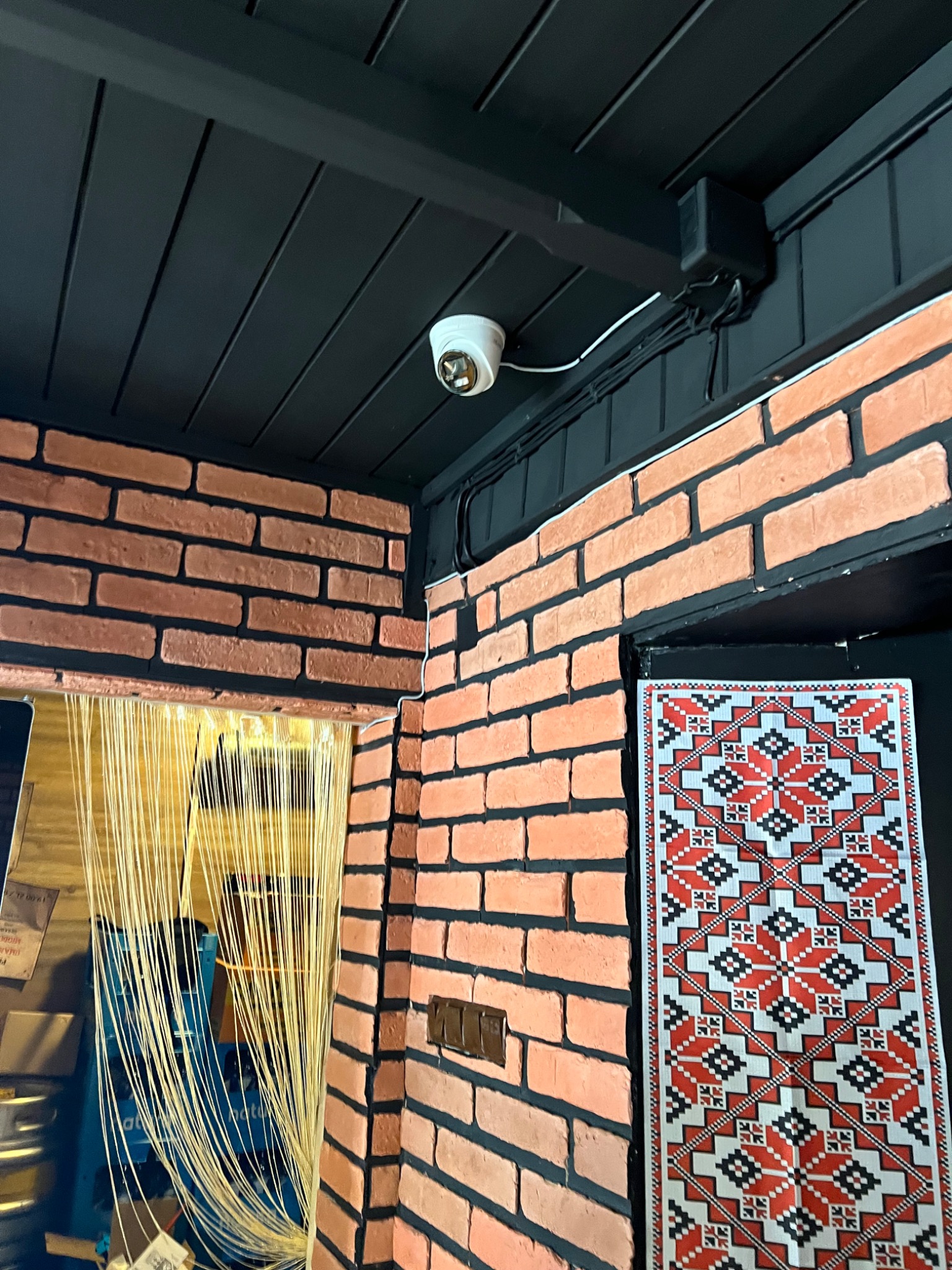 Kamera monitoringu kopułkowa zamontowana na czarnym suficie w pomieszczeniu z ceglanymi ścianami i dekoracyjnym elementem tekstylnym.