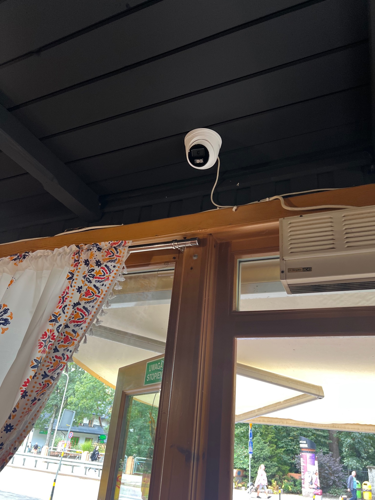 Kopułkowa kamera monitoringu zamontowana na czarnym, drewnianym suficie, widoczna nad ramą okienną z zasłoną i grzejnikiem. Widok na zewnątrz przez okno.