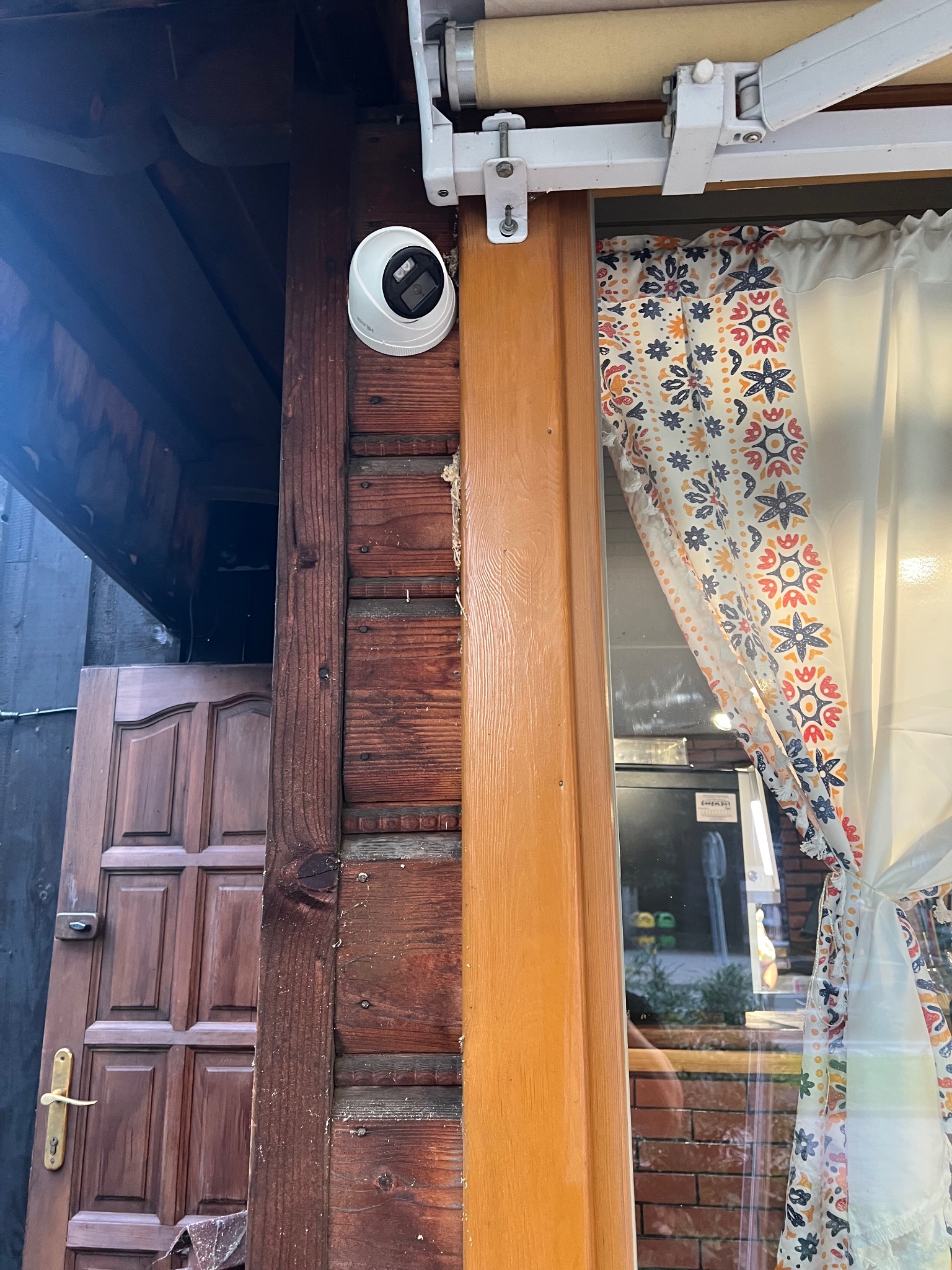 Kopułkowa kamera monitoringu zamontowana na drewnianej ścianie budynku, widoczna z boku, z elementami okna i zasłony w tle.