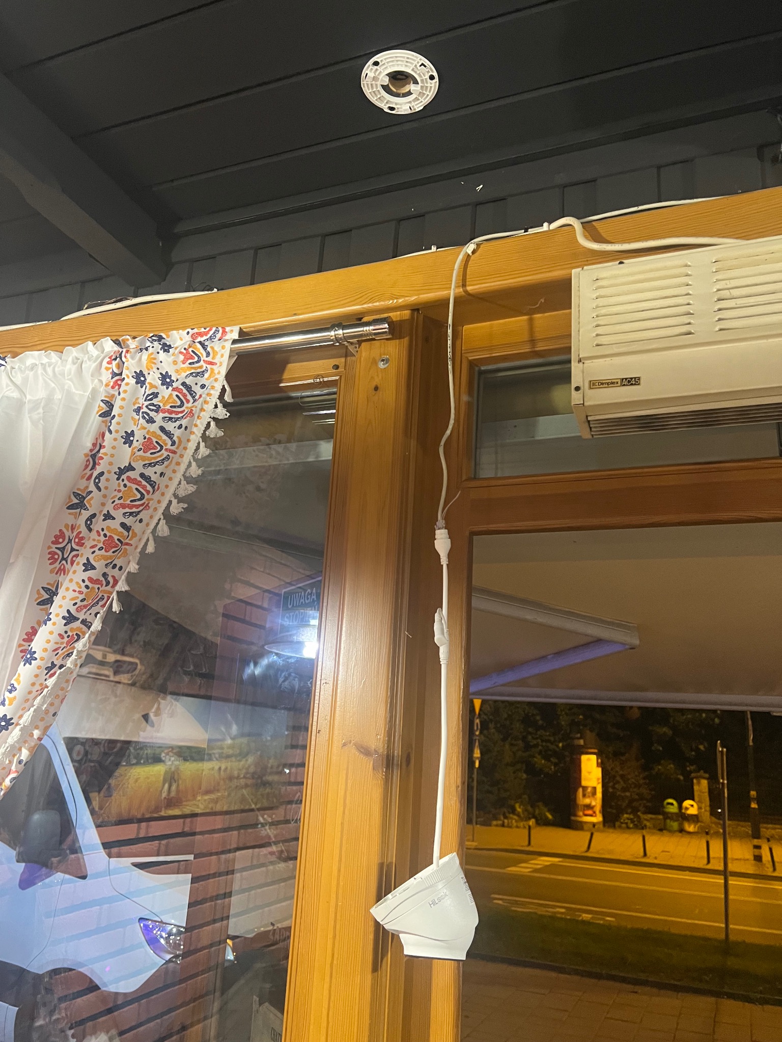 Wewnętrzny montaż kamery HiLook przy oknie z zasłoną i klimatyzacją Dimplex AC45. Widoczne przewody i uchwyt kamery. Zewnętrzne oświetlenie uliczne w tle.