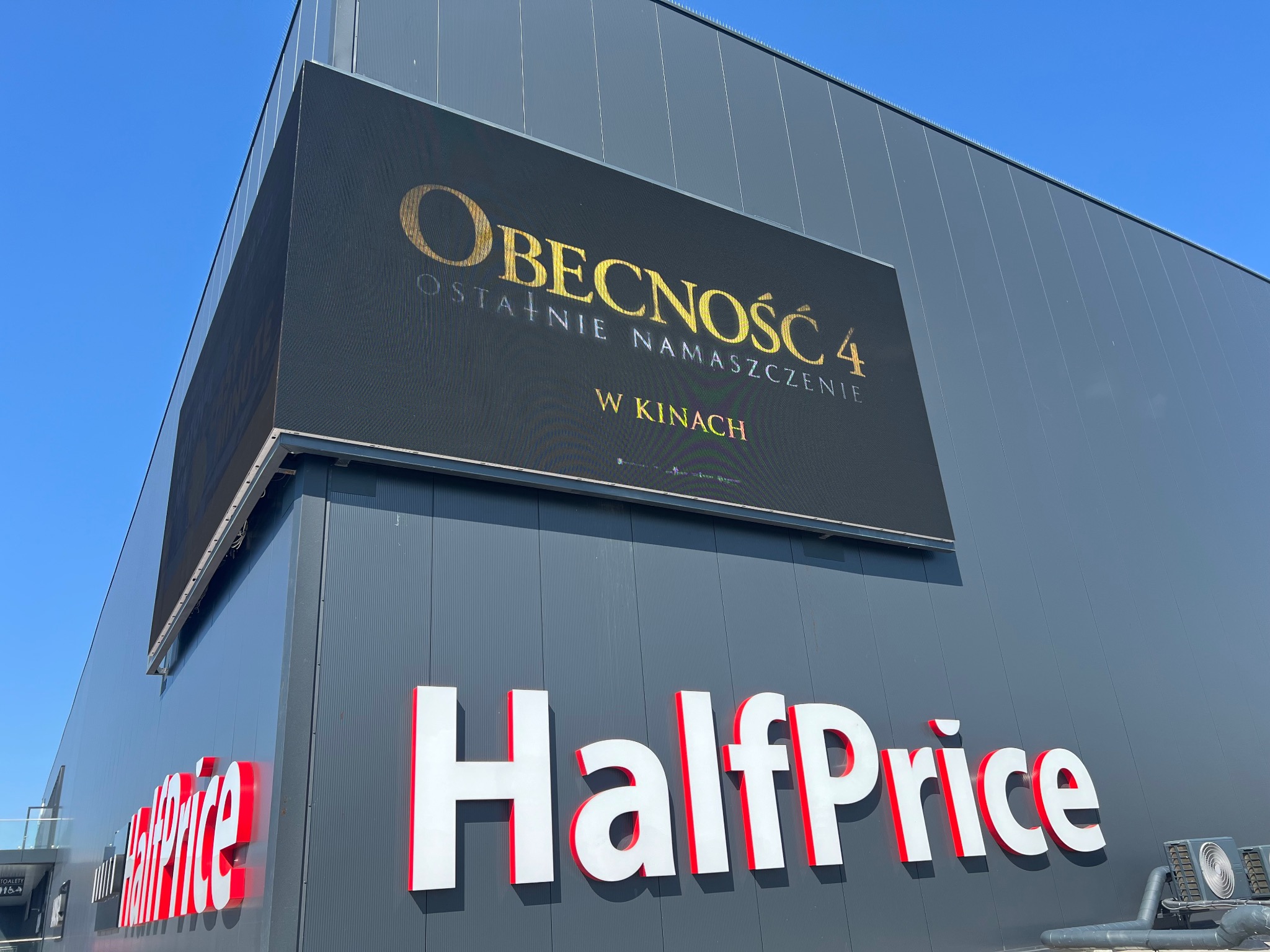 Elewacja budynku HalfPrice z LED ekranem wyświetlającym reklamę filmu 'Obecność 4'. Szary budynek, biało-czerwone logo. Błękitne niebo.