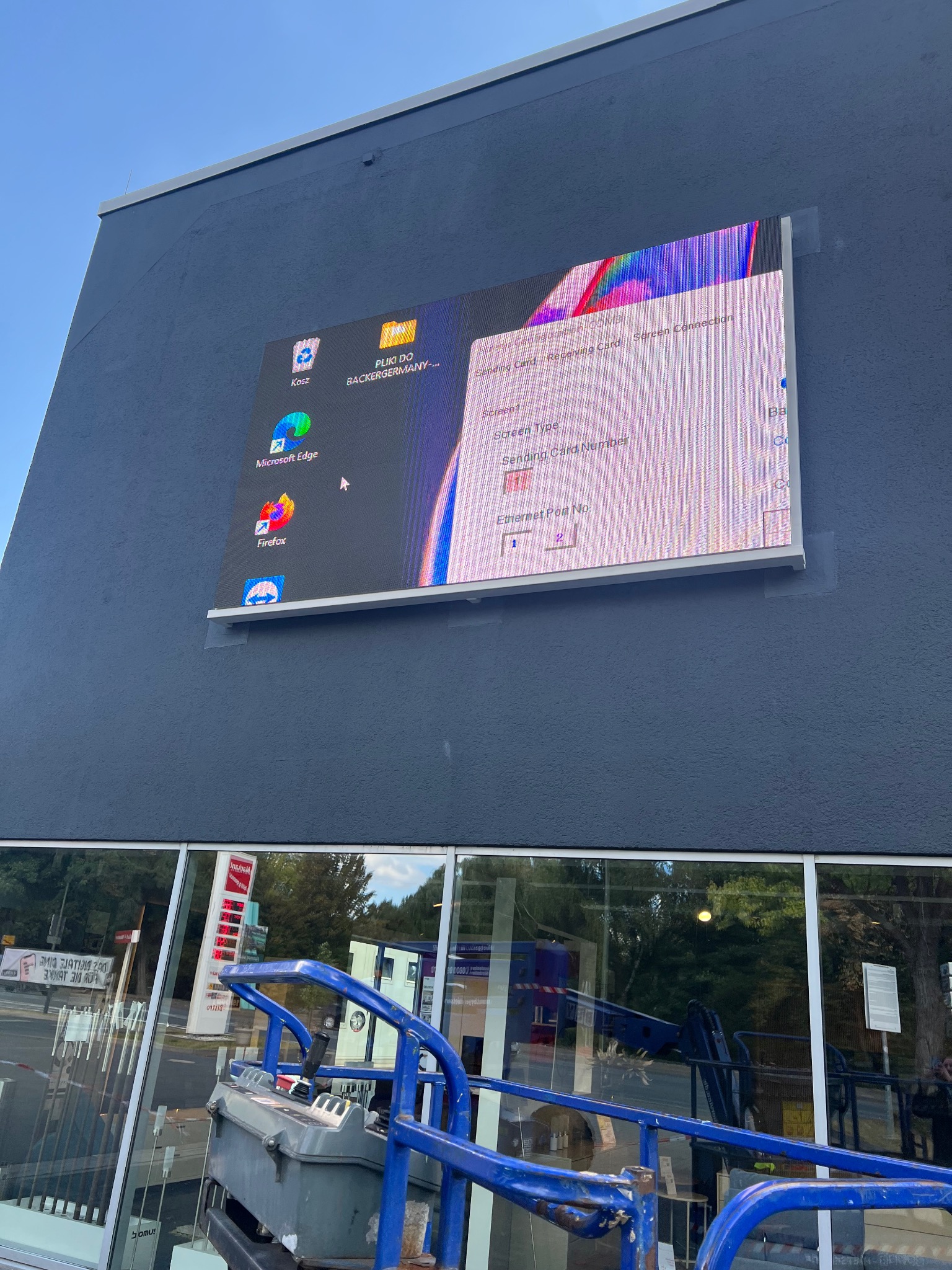 Instalacja zewnętrznego ekranu LED na elewacji budynku, widoczny pulpit systemu operacyjnego z ikonami i ustawieniami. Prace montażowe z użyciem podnośnika koszowego.