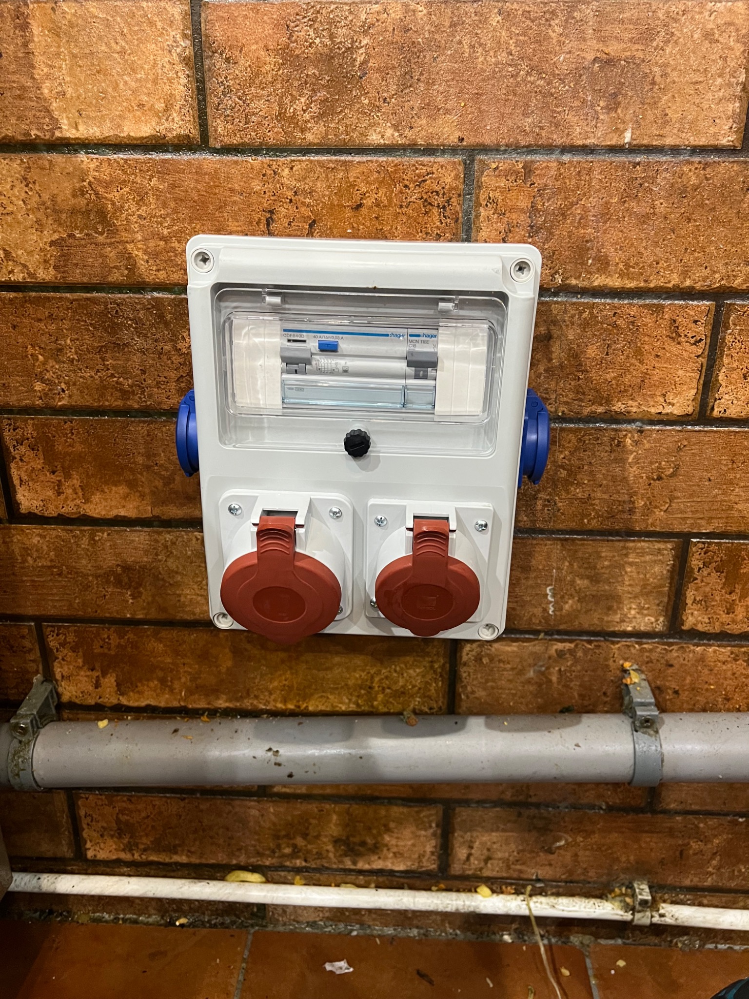 Biała skrzynka elektryczna z dwoma gniazdami przemysłowymi na ceglanej ścianie, widoczne zabezpieczenia wewnątrz skrzynki i szara rura instalacyjna poniżej.