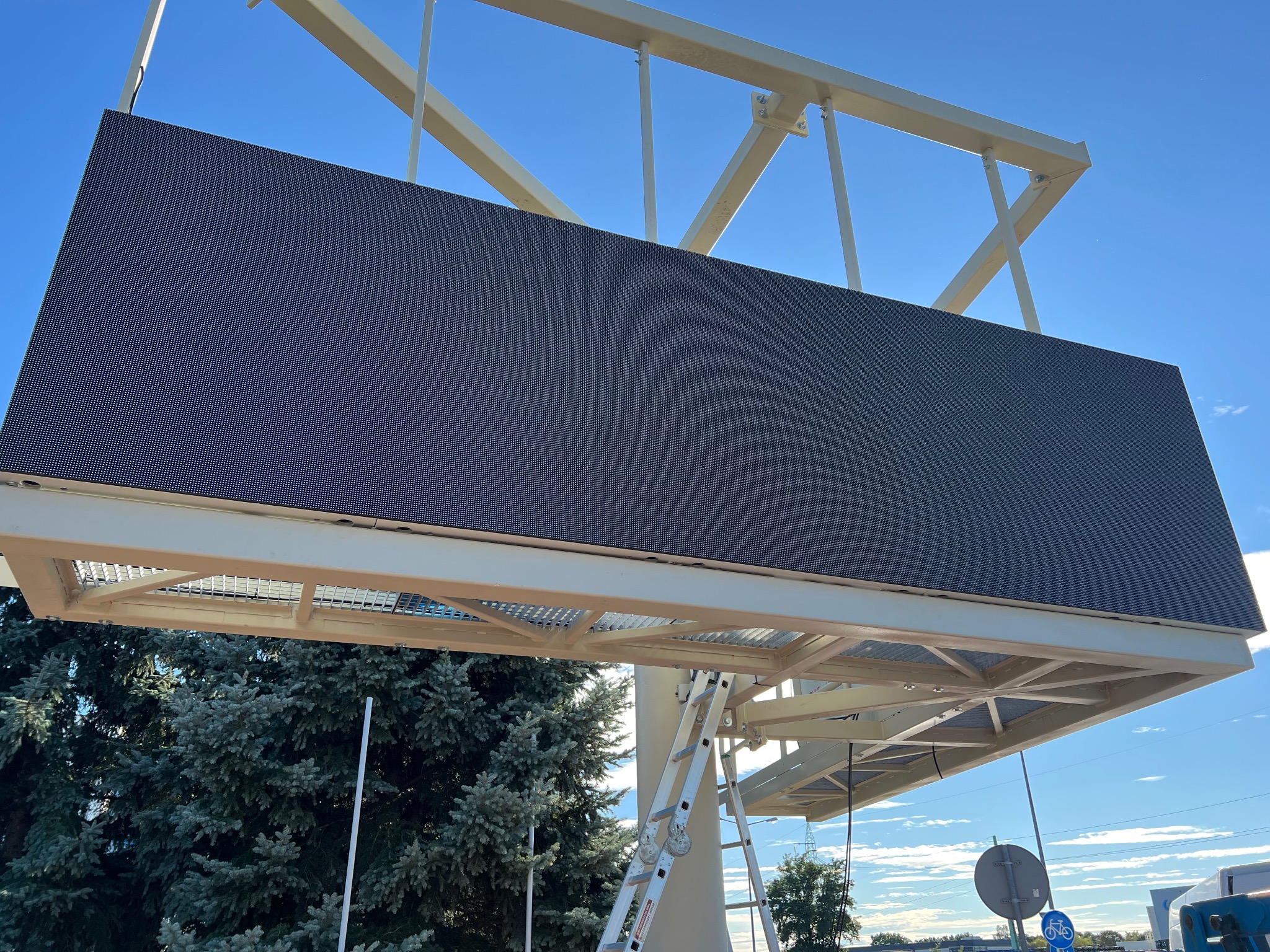 Duży ekran LED na metalowej konstrukcji, widoczny z dołu, na tle błękitnego nieba. Drabina oparta o konstrukcję. Widoczne drzewa i znaki drogowe.